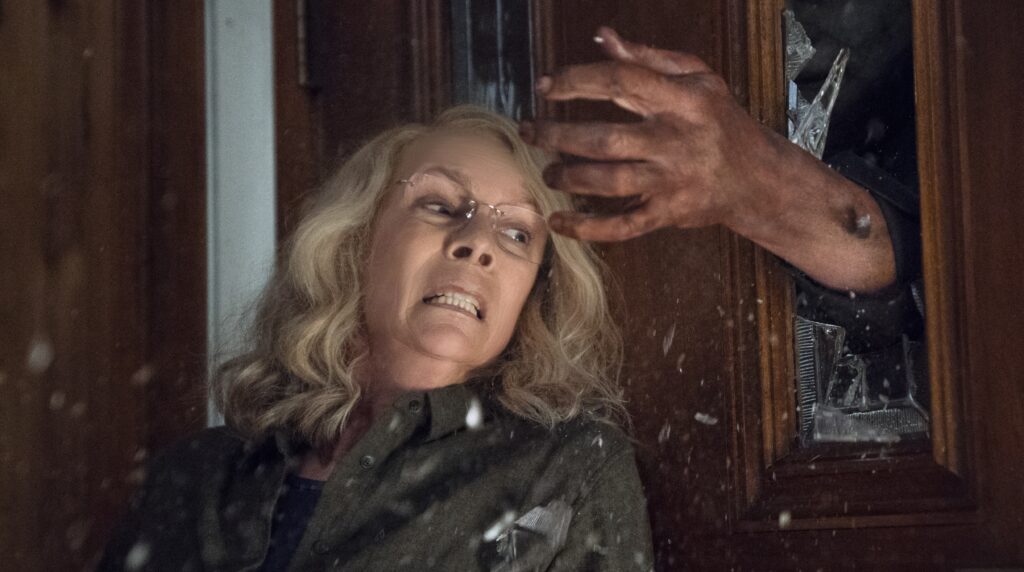 Halloween (2018) Screencap with Jamie Lee Curits as Laurie as a Hand Reaches Through Broken Glass to Get Her