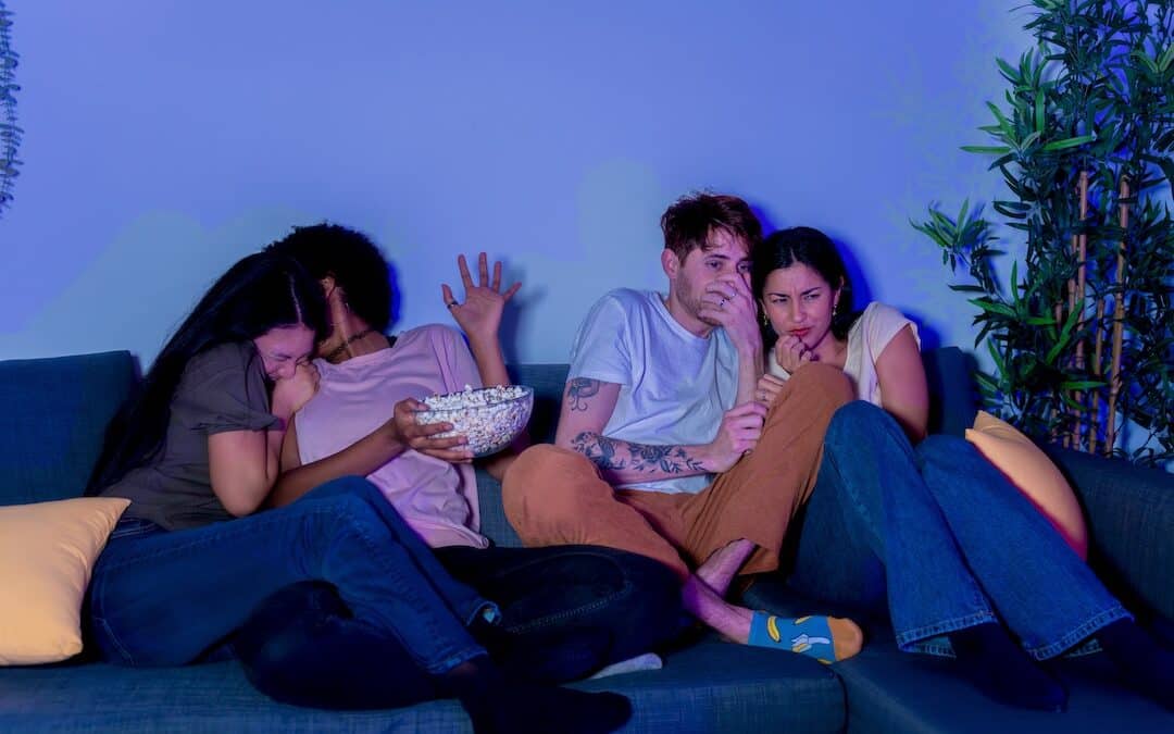 Terrified friends huddled together on a couch watching a scary movie at night.