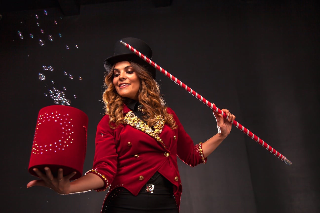 Female magician with box and magic cane makes with soap bubbles show, an illusionist in theatrical clothes and hat. Woman actress in stage costume. Concept of theatrical performance and fun show Christmas circus