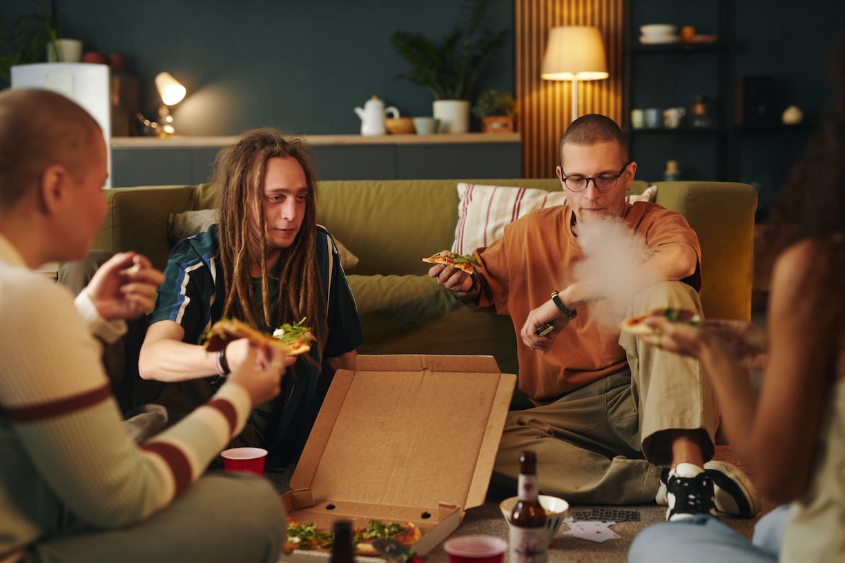 Group of friends sitting on couch, sharing pizza and relaxing together. Warm, cozy living room setting with pizza box