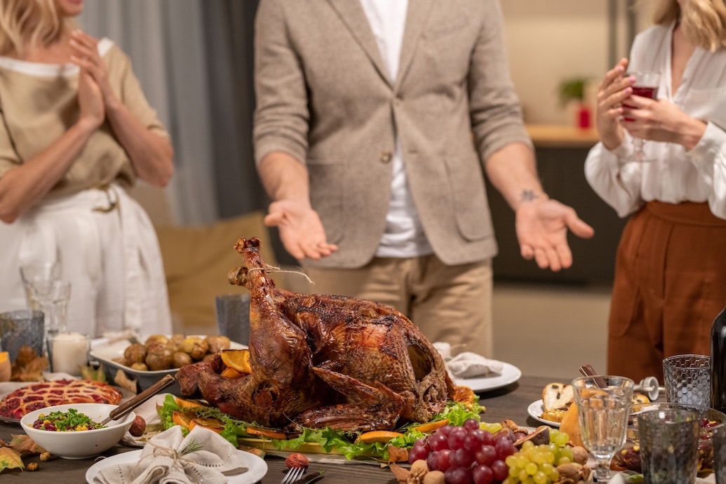 Homemade roasted turkey among fresh fruits, salad and desserts on served table on background of three family members having discussion