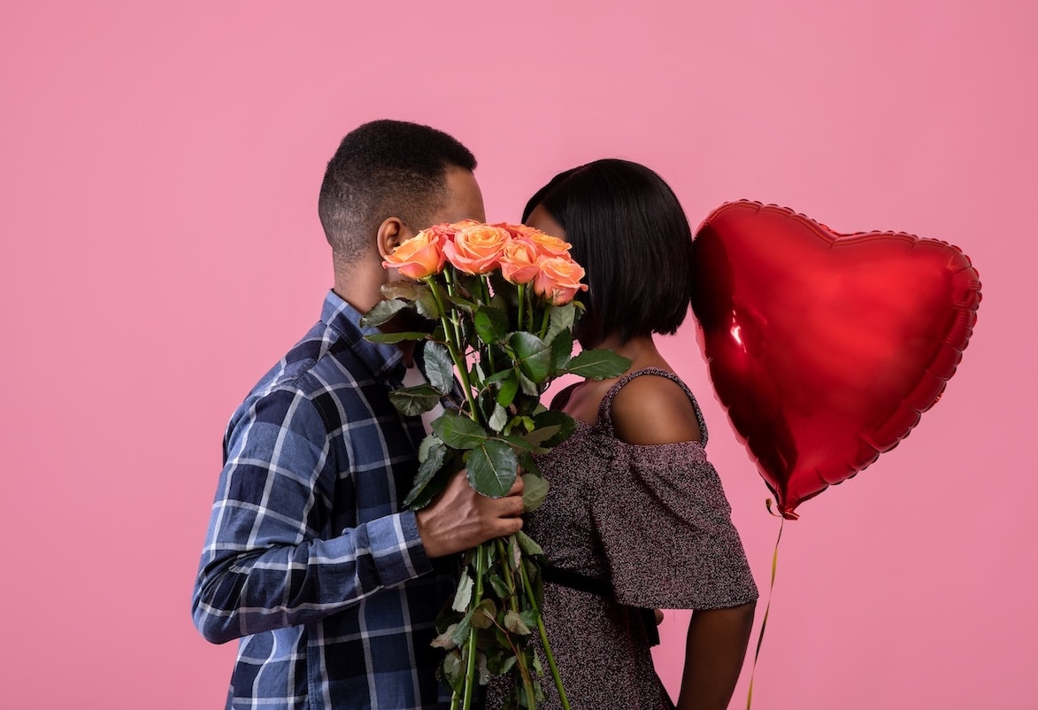 Couple on a Pink Backdrop. One is Holding a Bouquet of Roses, the Other is Holding a Heart Shaped Balloon