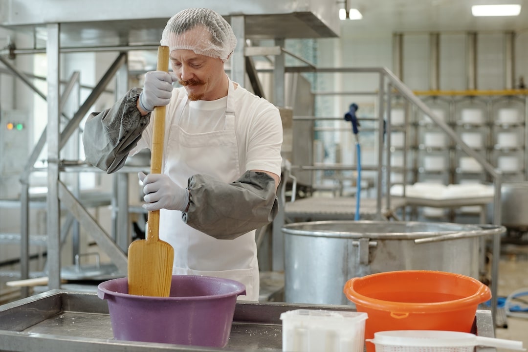 Man wearing protective gear stirring yellow mixture in large container in industrial kitchen surrounded by various equipment and containers with focus on his work