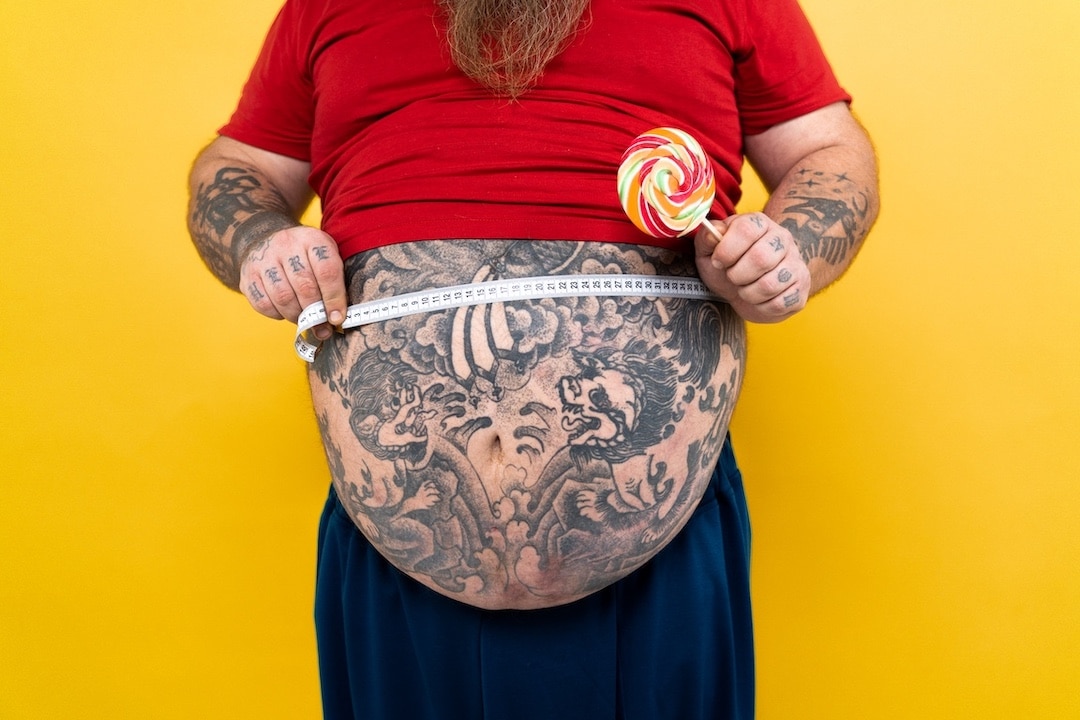 Fat guy with tattooed belly holding lolly pop and measuring stomach with tape measure