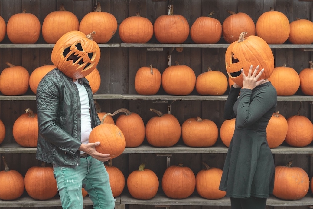 Two People with Jack o' Lanterns on Their Heads in Front of a Wall Lined with Pumpkins